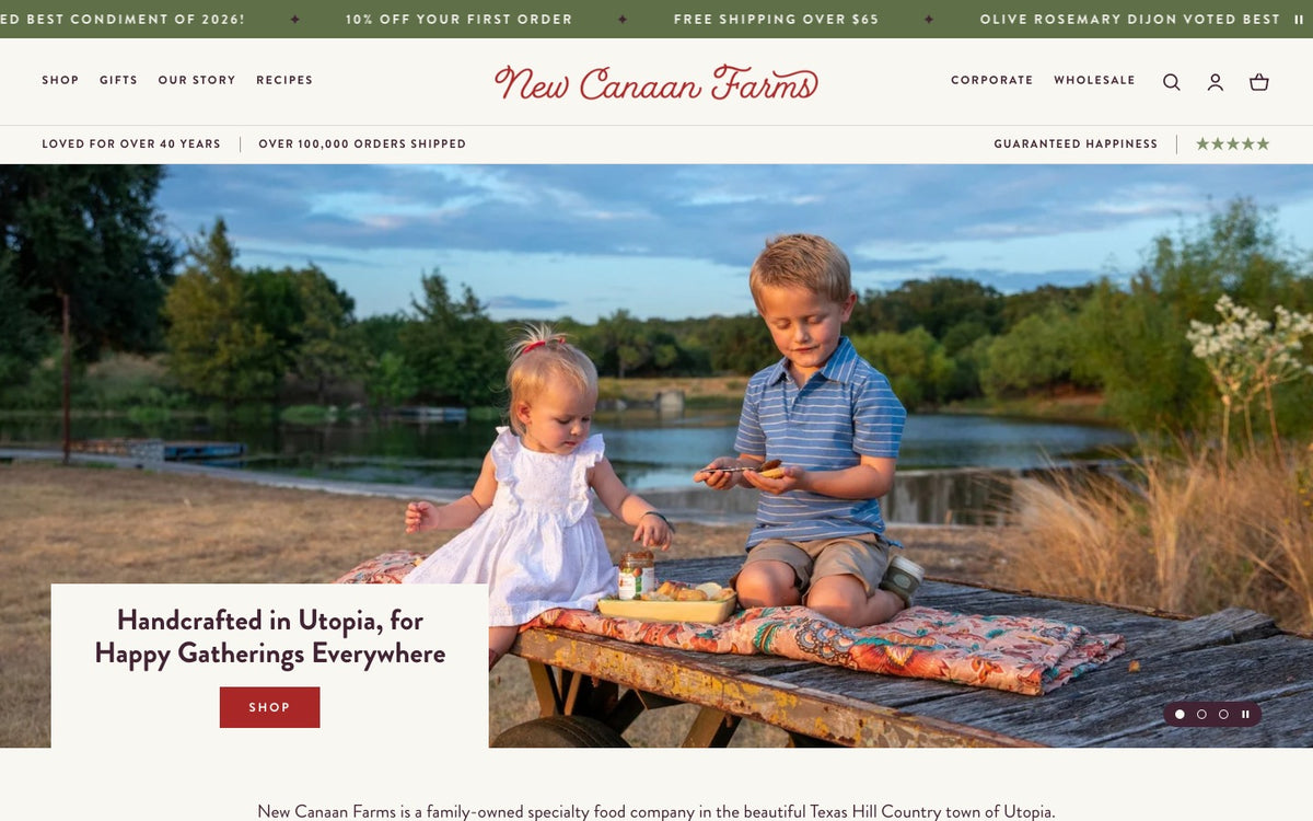The new homepage. A green announcement ticker and a trust bar sit around a centered script logo, above a photo of two children sharing a jar of jam at a creek-side picnic in Utopia. A cream panel on the photo's bottom edge holds the headline Handcrafted in Utopia, for Happy Gatherings Everywhere and a red Shop button.